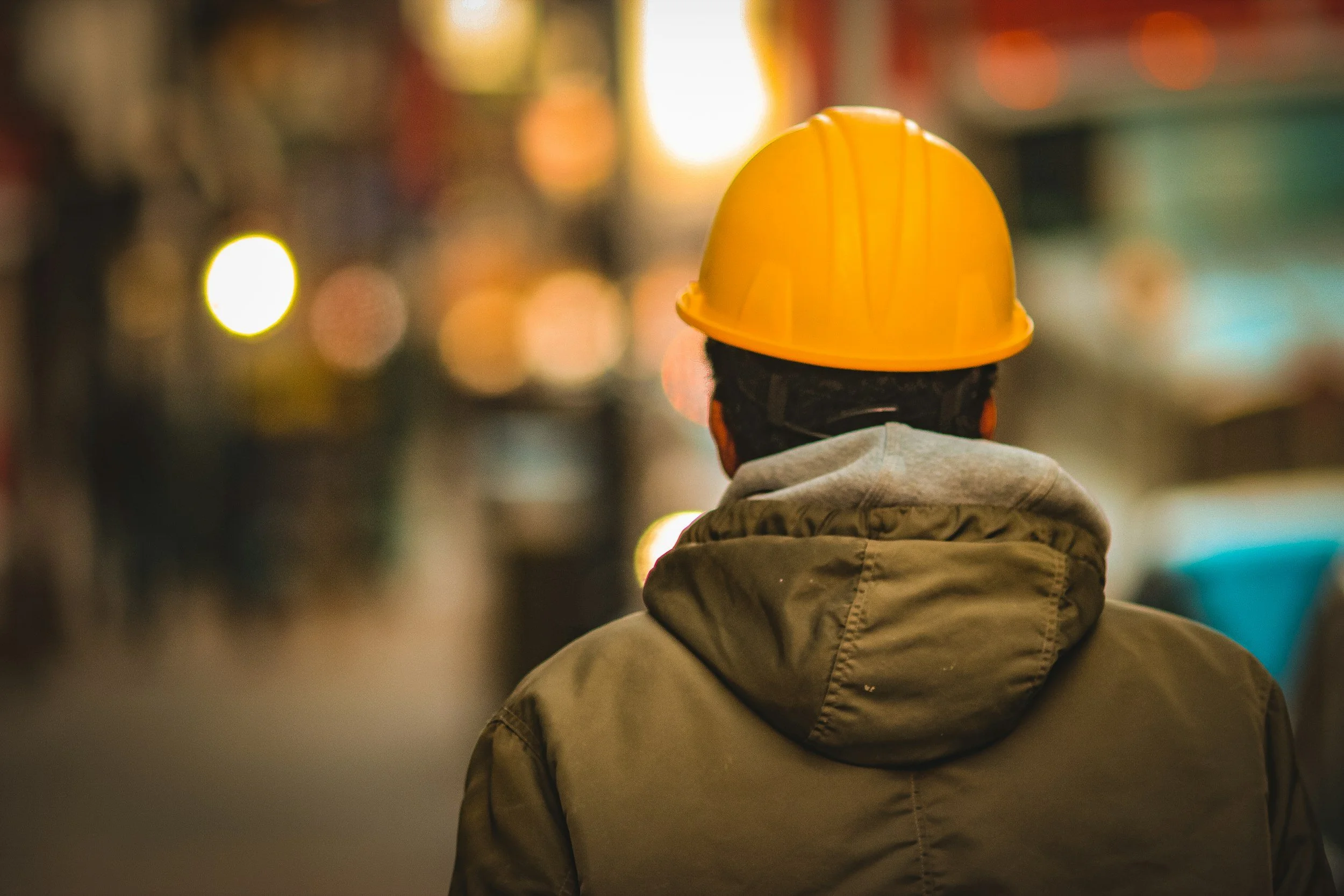 Person wearing a yellow hard hat and jacket, standing outdoors with blurred lights in the background.